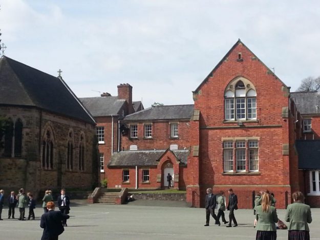 Heinemann_1 Mehrere Schüler und Schülerinnen vor einem roten Gebäude (Internat) und einer Kirche.