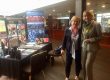 Zwei blonde Frauen stehen neben einem Tisch mit Plakaten und Werbematerial und lächeln.