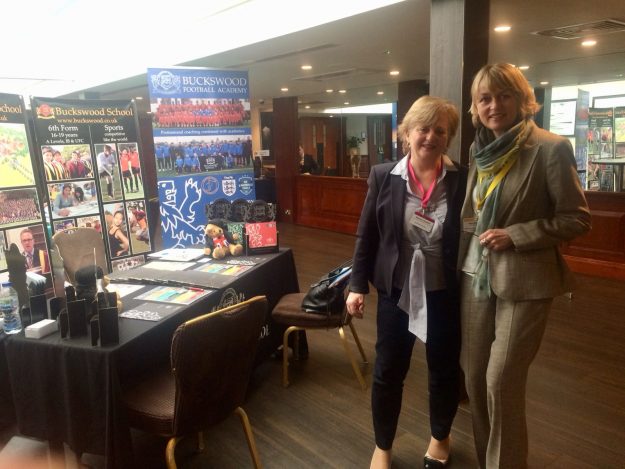 image1 Zwei blonde Frauen stehen neben einem Tisch mit Plakaten und Werbematerial und lächeln.