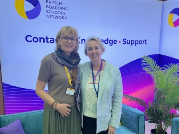 Petra Kirschke & Suzanne Rowse_640x480 Zwei Frauen stehen Arm in Arm vor einem Sofa. Dahinter ist ein Banner des British Boarding School Networks.